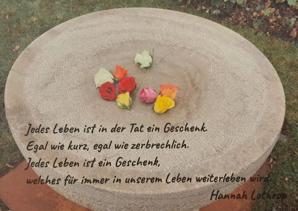 Stein mit Rosenköpfen darauf und dem Spruch: Jedes Leben ist in der Tat ein Geschenk. Egal wie kurz, egal wie zerbrechlich. Jedes Leben ist ein Geschenk, welches für immer in unserem Leben weiterleben wird. (von Hannah Lothrop)